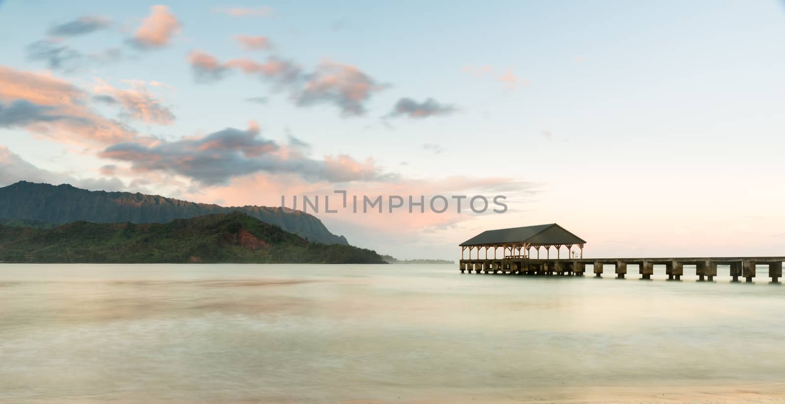 Rising sun illuminates the peaks of Na Pali mountains over the calm bay and Hanalei Pier in long exposure photo