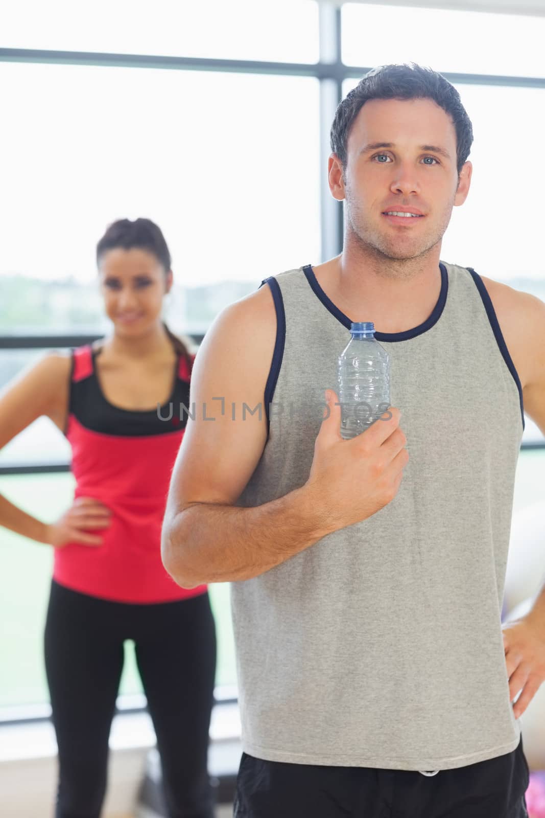 Portrait of a fit young man holding water bottle with friend in background in a bright exercise room