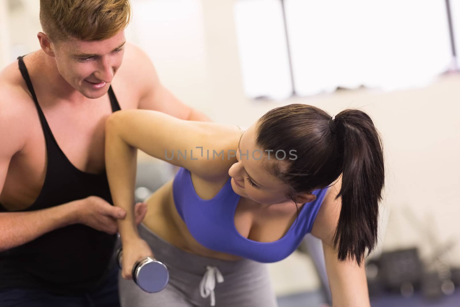 View of a male trainer helping young woman with dumbbell in the gym