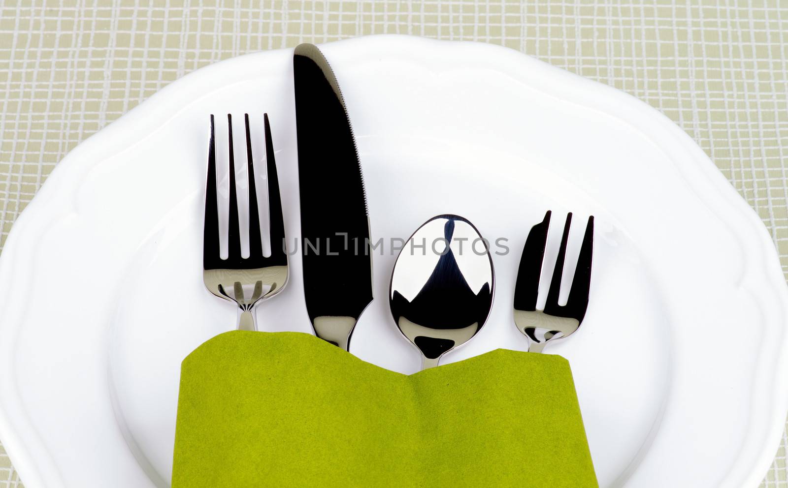 Elegant Table Setting with Fork, Table Knife, Spoon and Dessert Fork into Green Napkin Decorated on White Plate closeup on Checkered background 