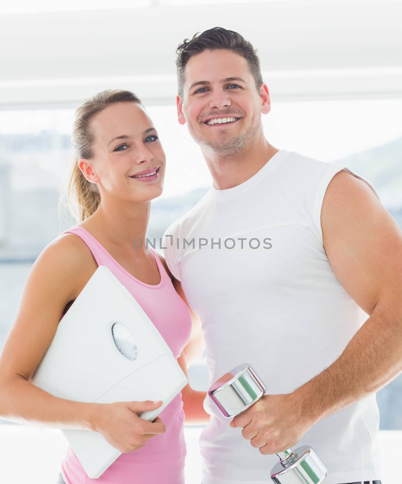 Portrait of a fit couple holding dumbbell and weighing scale in bright exercise room