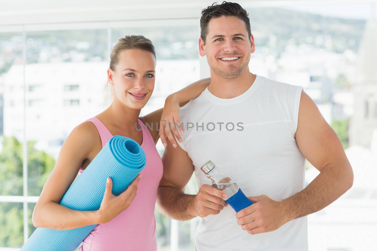 Portrait of a fit couple holding water bottle and exercise mat in bright exercise room