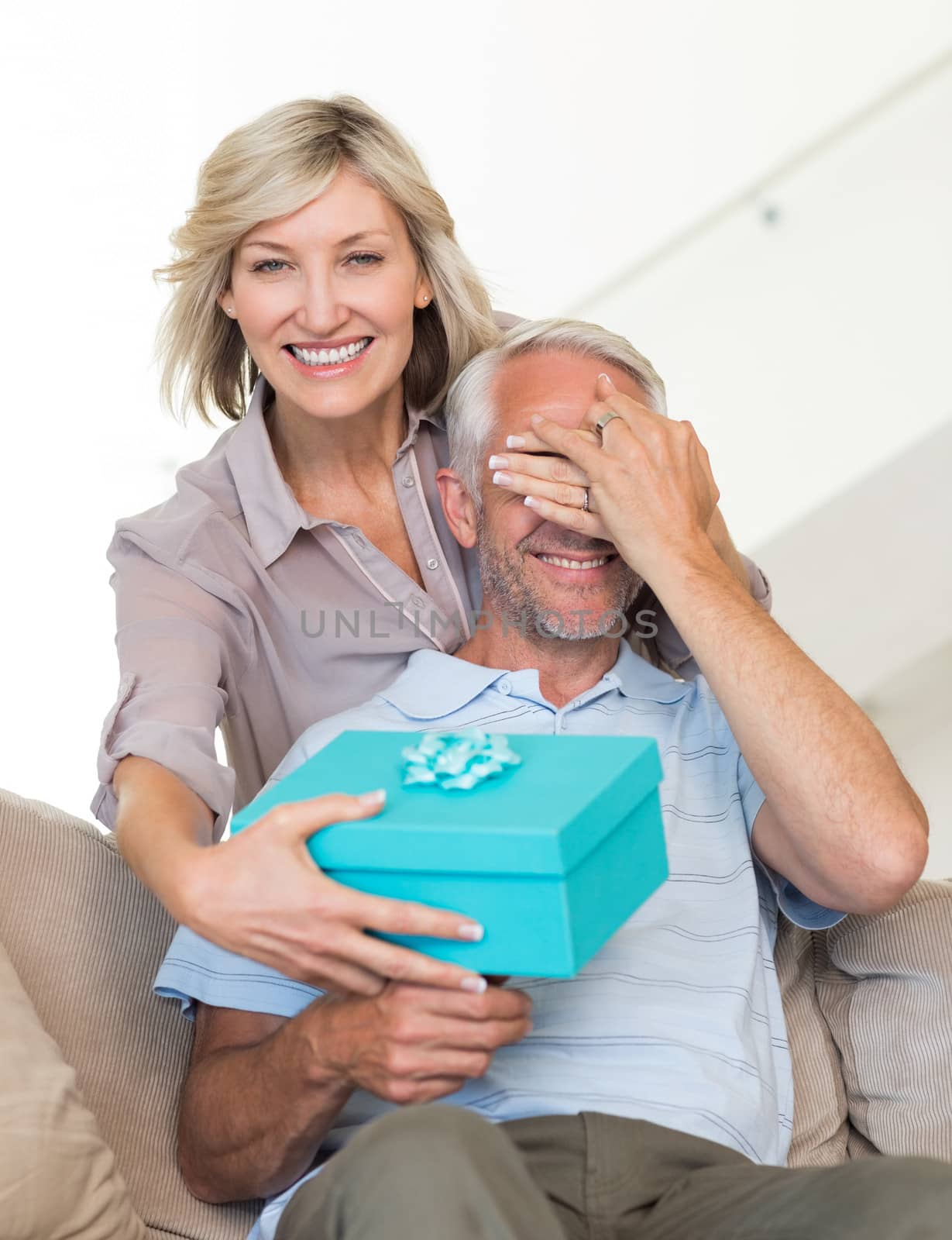 Portrait of a smiling woman surprising mature man with a gift on sofa at home