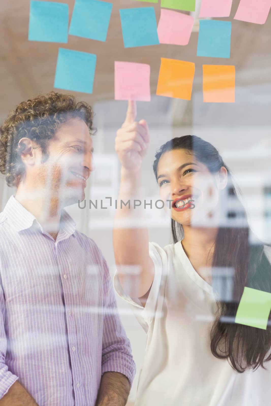 Happy businessman and businesswoman discussing over adhesive notes on glass wall in office