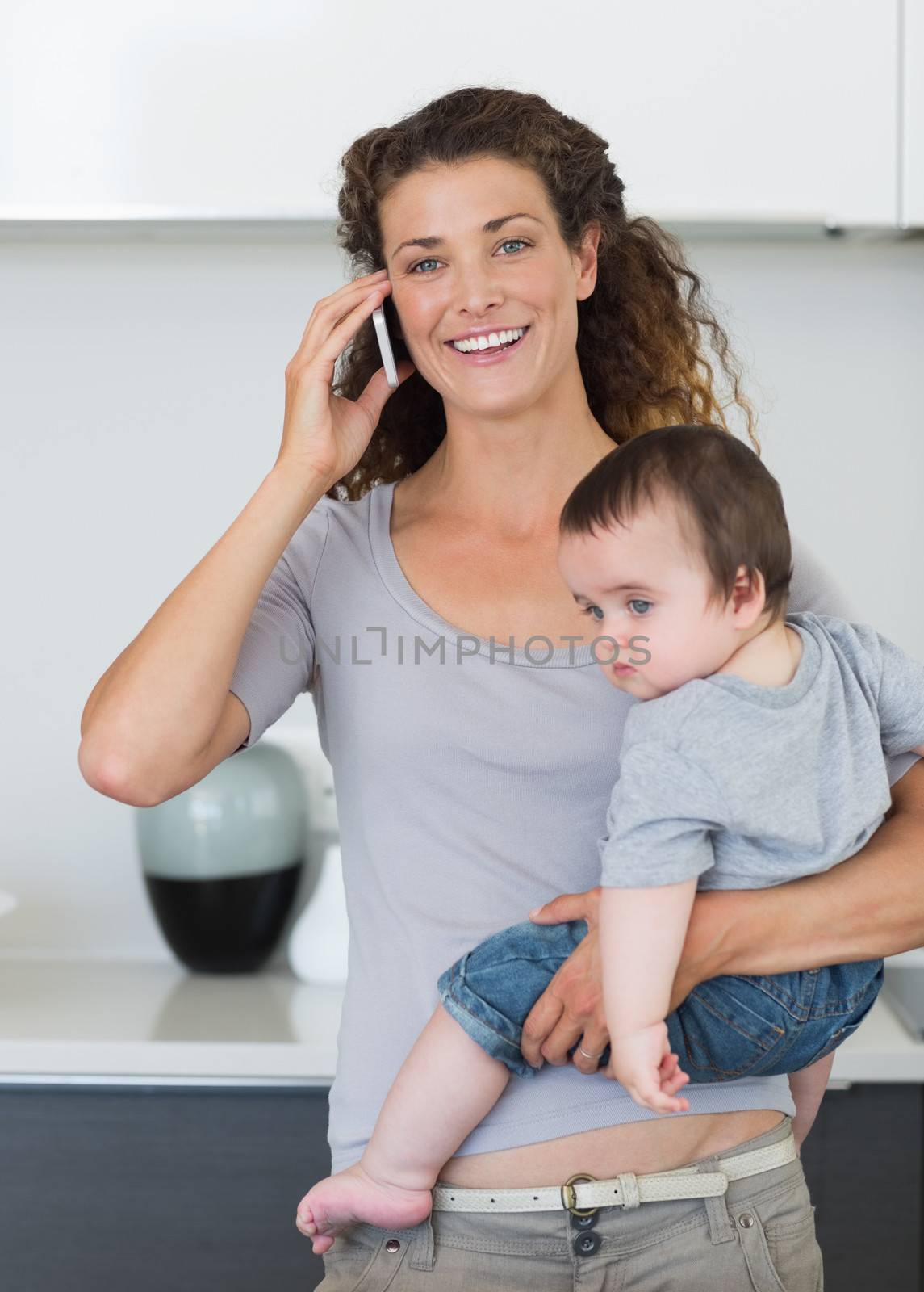Happy woman answering smartphone while carrying baby boy in kitchen