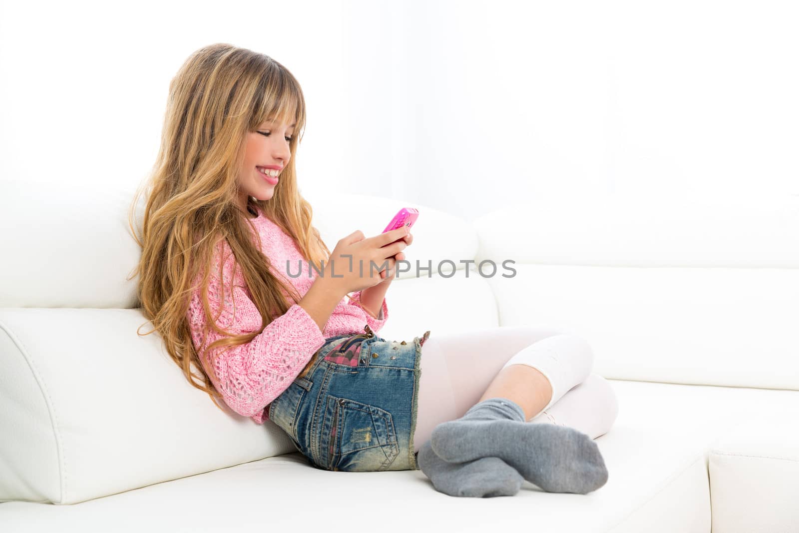 Blond kid girl playing fun with mobile smartphone on white sofa byod