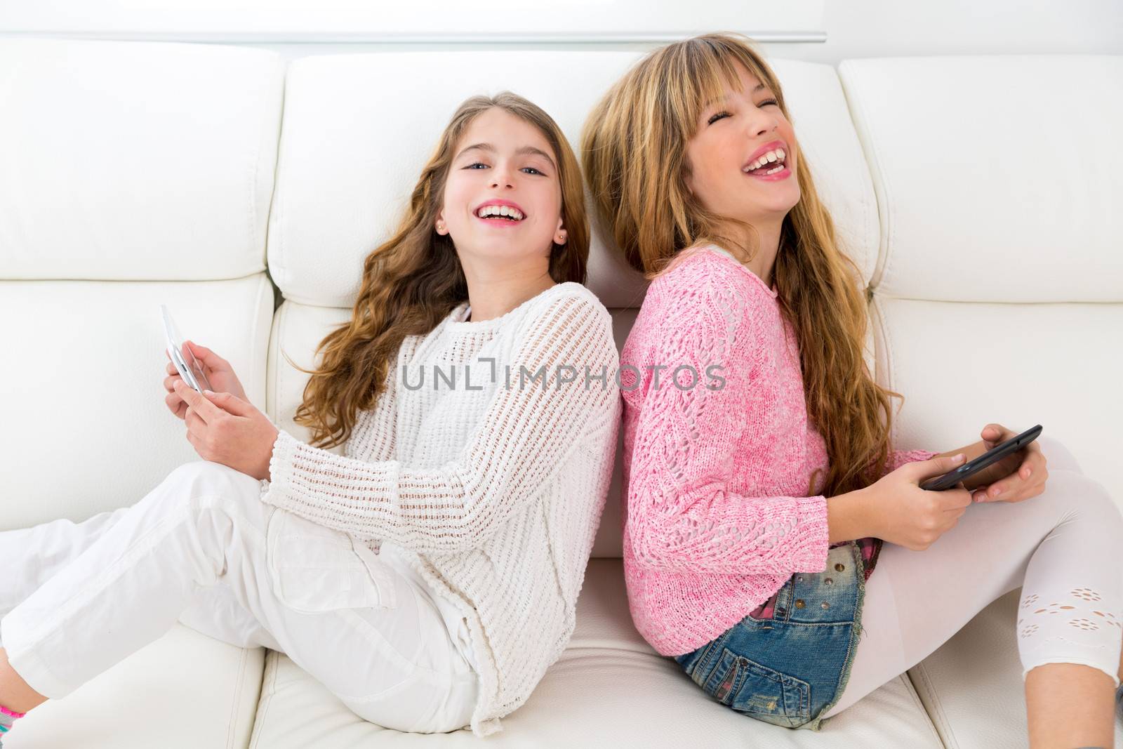 Children friends kid girls having fun playing back to back with tablet pc on white sofa