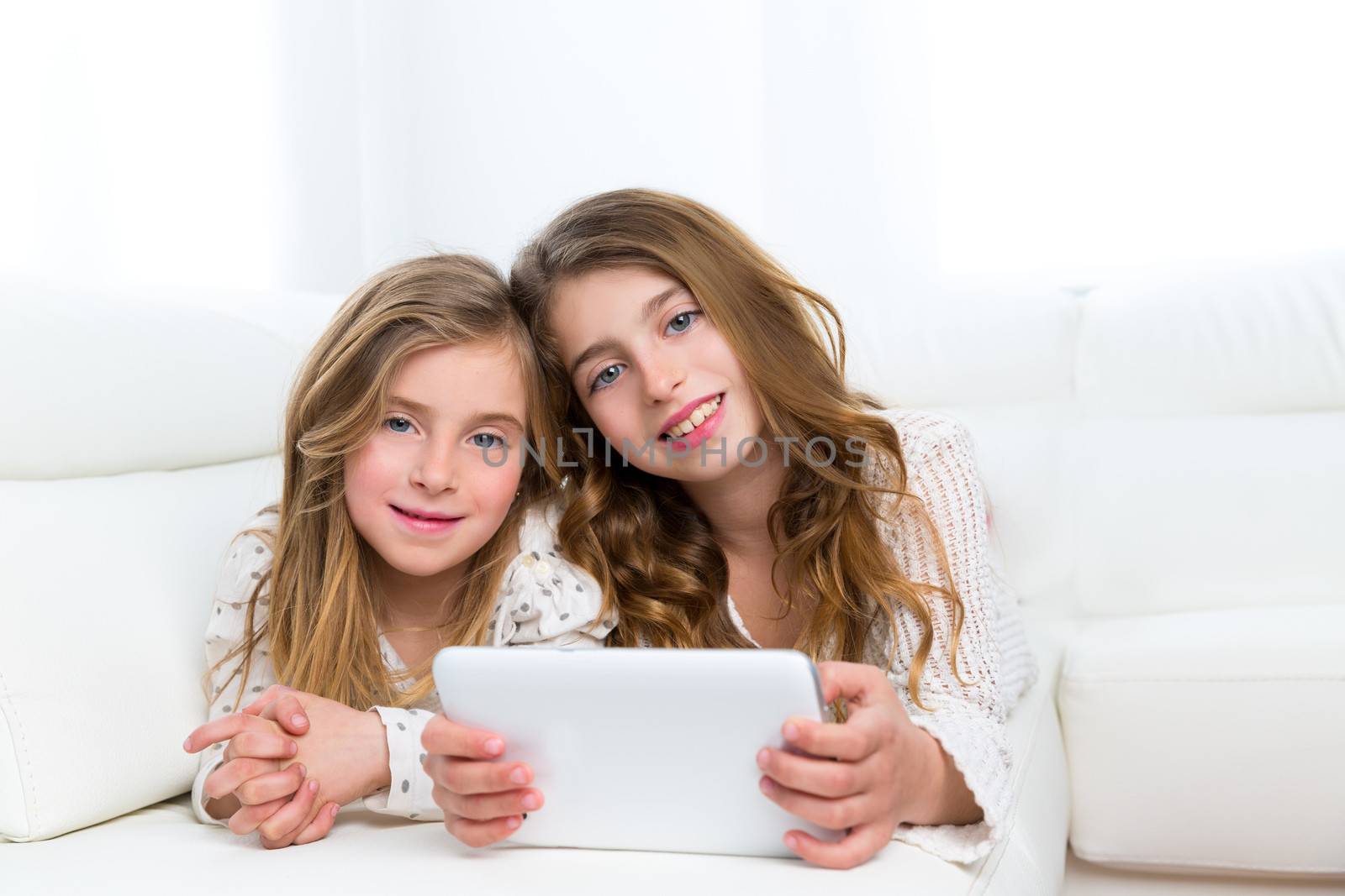 Children sister friends kid girls playing together with tablet pc lying on white sofa