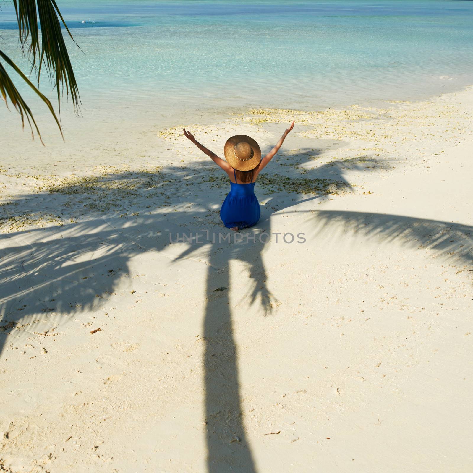 Woman in blue dress on a tropical beach at Maldives sitting in palm tree shadow