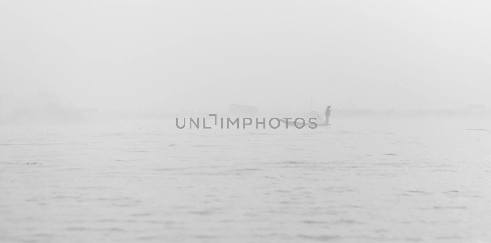 Foggy morning weather on a lake with silhouette of fisherman in small wooden boat 