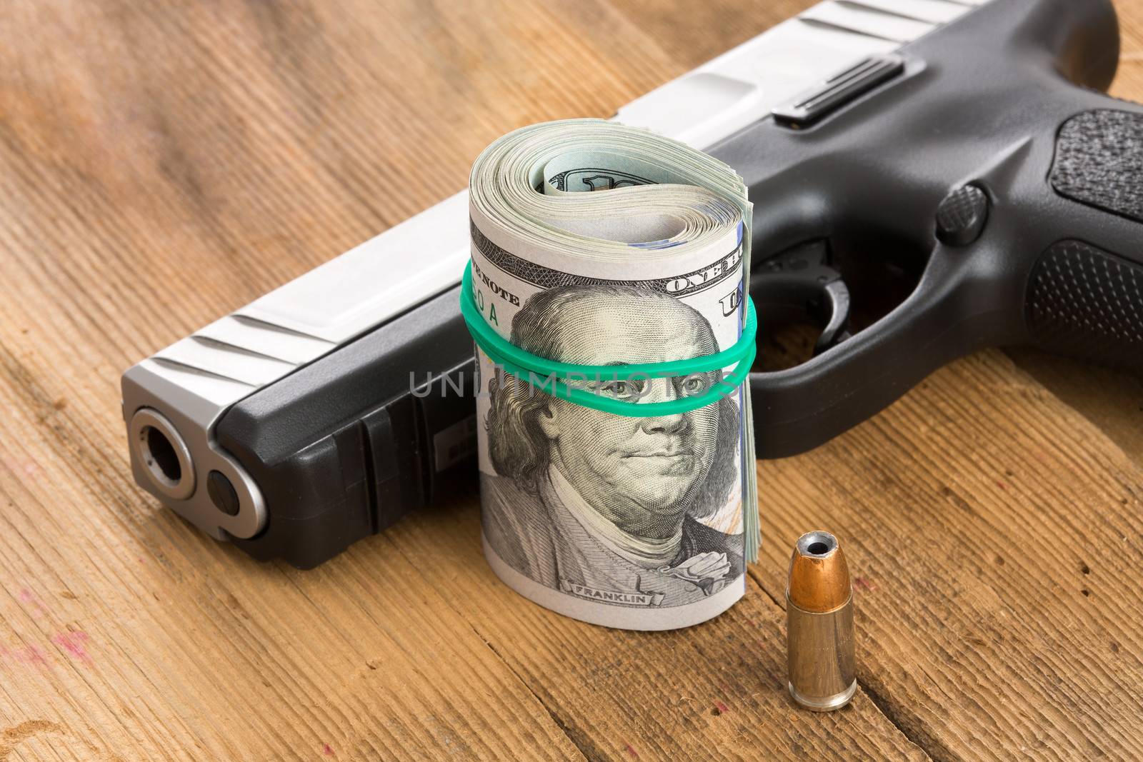 Handgun with a roll of 100 dollar bills and a single bullet lying on a wooden surface with the barrel and muzzle towards the camera conceptual of crime, a payoff, gun for hire or protection