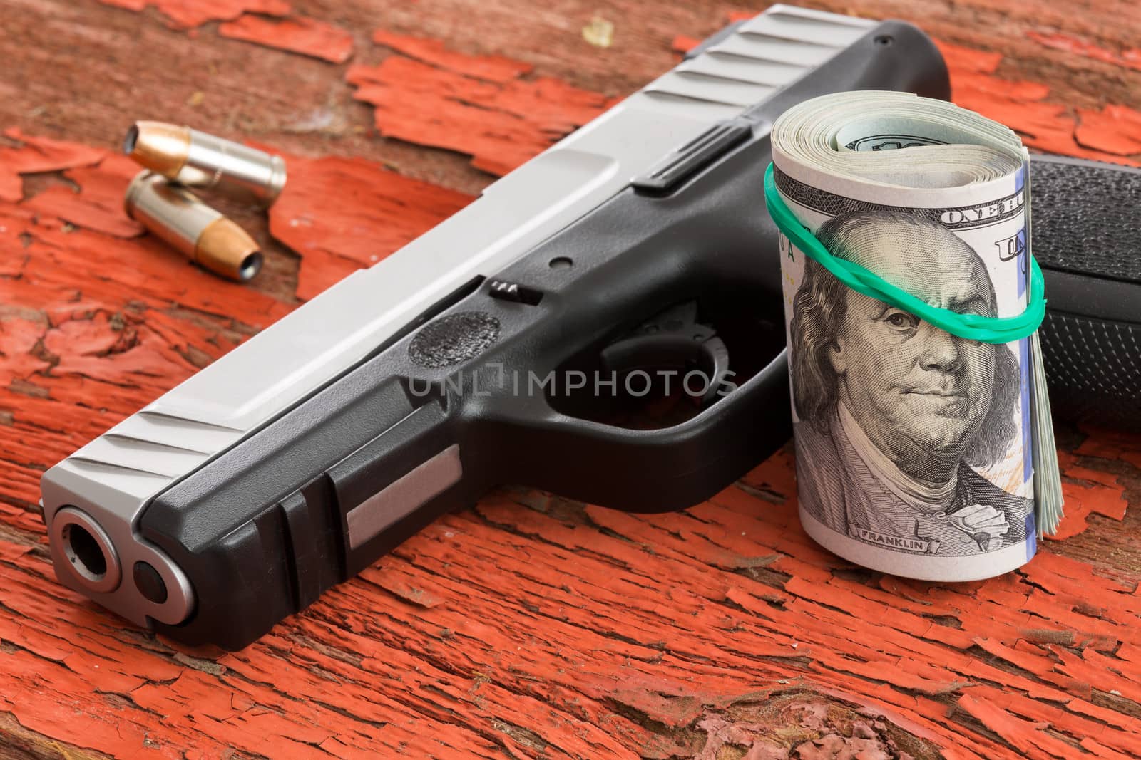 Gun with a roll of 100 dollar banknotes and two bullets lying on a grungy wooden surface with red peeling paint conceptual of crime, robbery, coercion, bribe or mob protection money