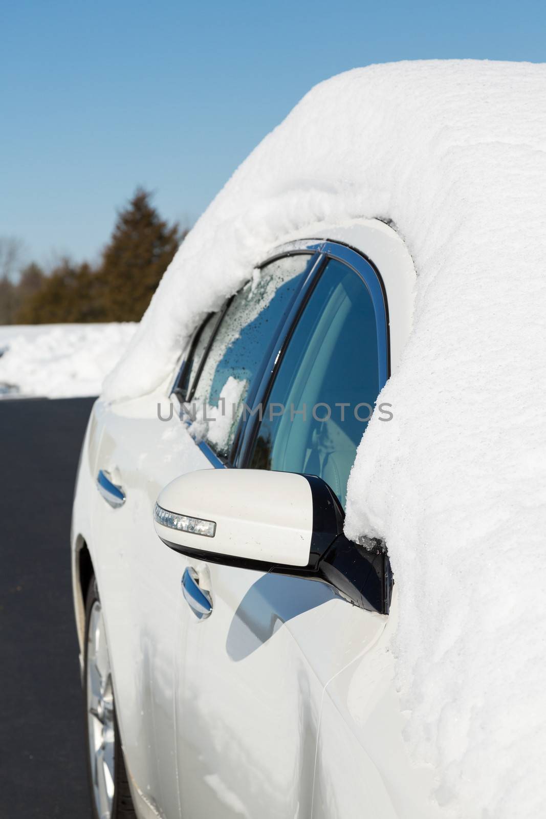 Deep snow piled high on top of white four door sedan car in rural setting with view down side of car and drive