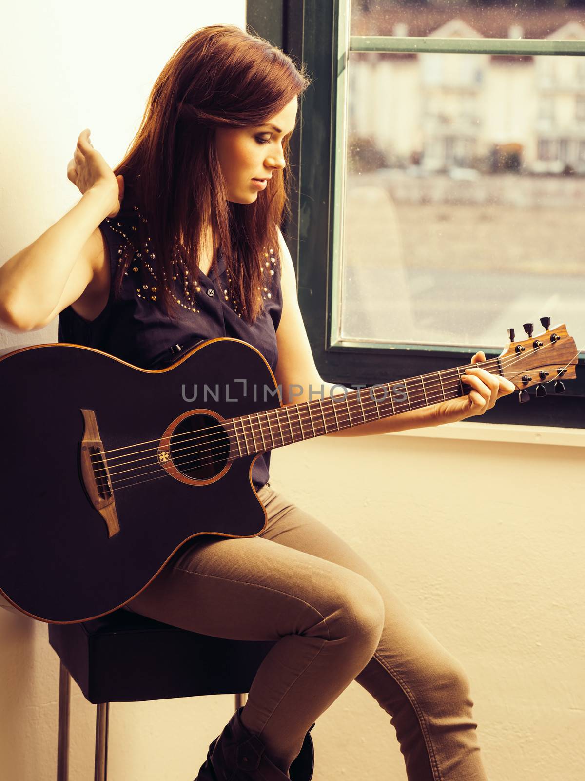 Photo of a woman playing an acoustic guitar while sitting by a window.