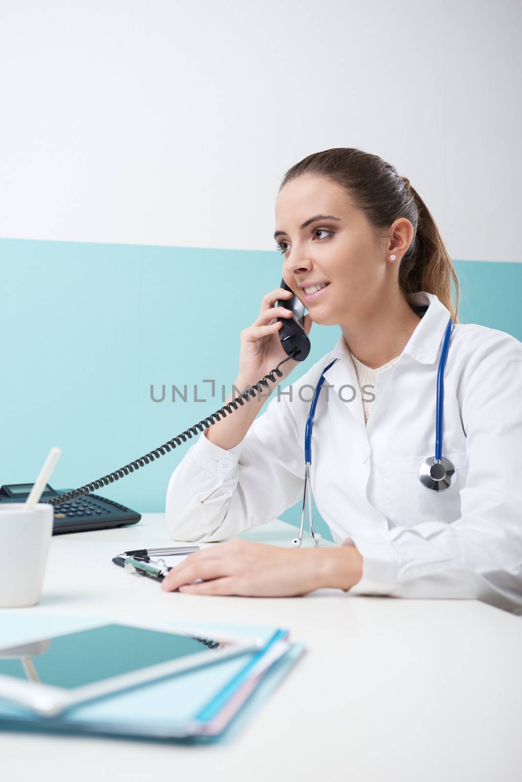 Young female doctor sitting at her desk and talking on the phone