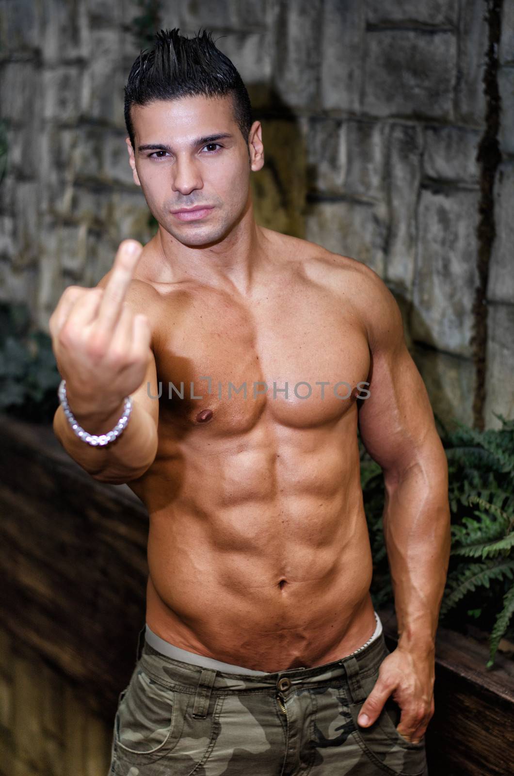 Muscular, shirtless young man doing 'screw you' sign with middle finger looking at camera