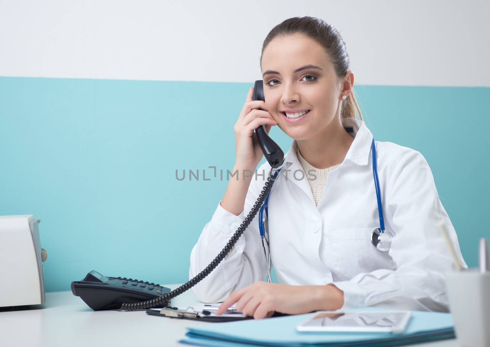 Young female doctor sitting at her desk and talking on the phone