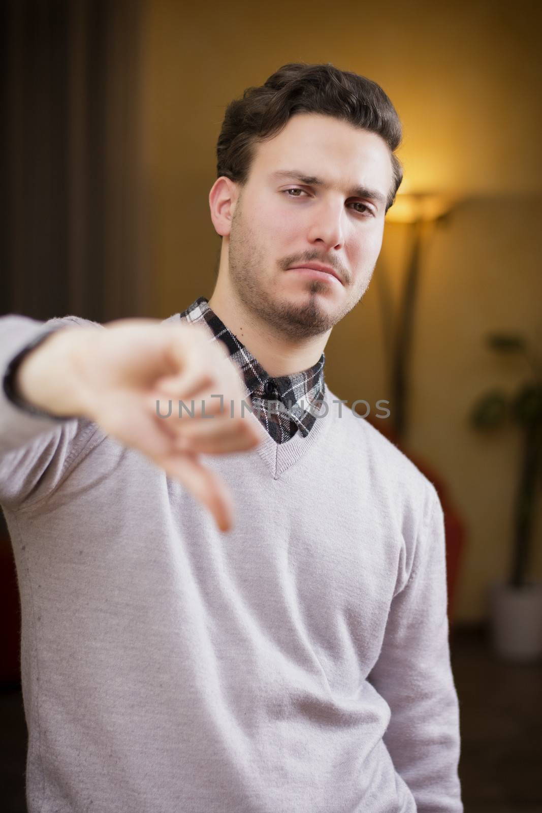 Disappointed or displeased young man doing thumb down sign. Indoors shot inside a house