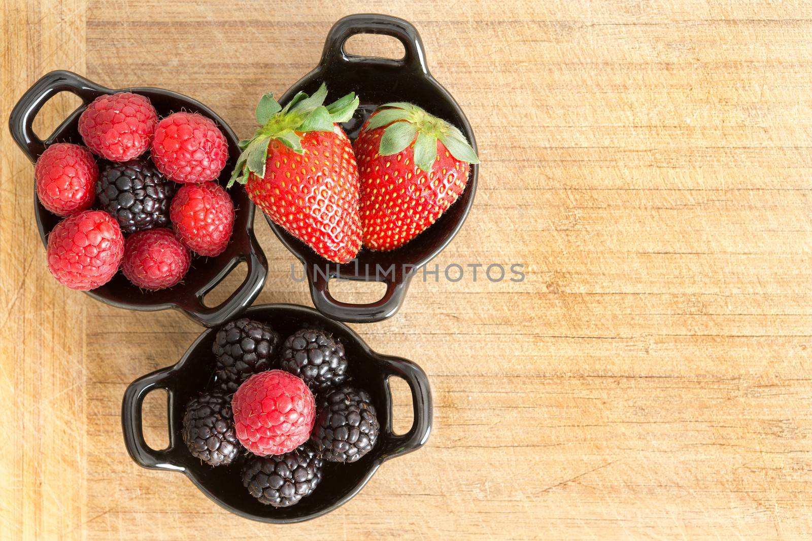 Overhead view of three ceramic ramekins full of healthy ripe fresh mixed berries including strawberries, blackberries and raspberries on a wooden surface with copyspace