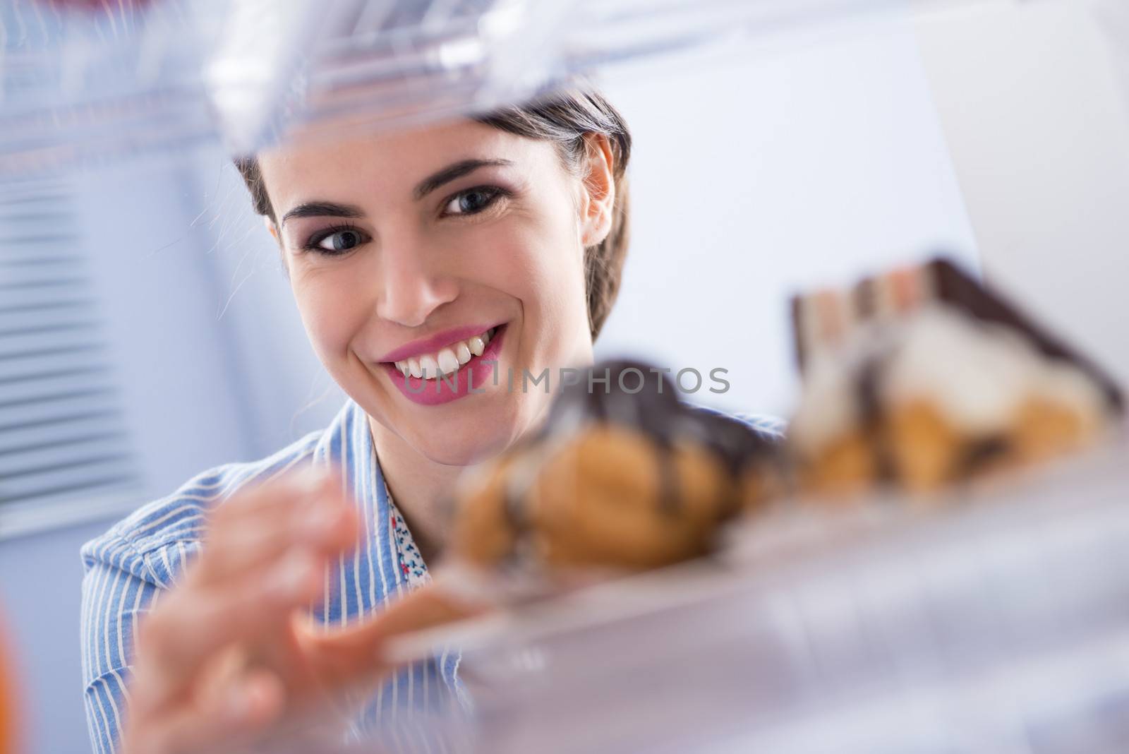 Young woman smiling and taking pastry with chocolate topping from fridge.