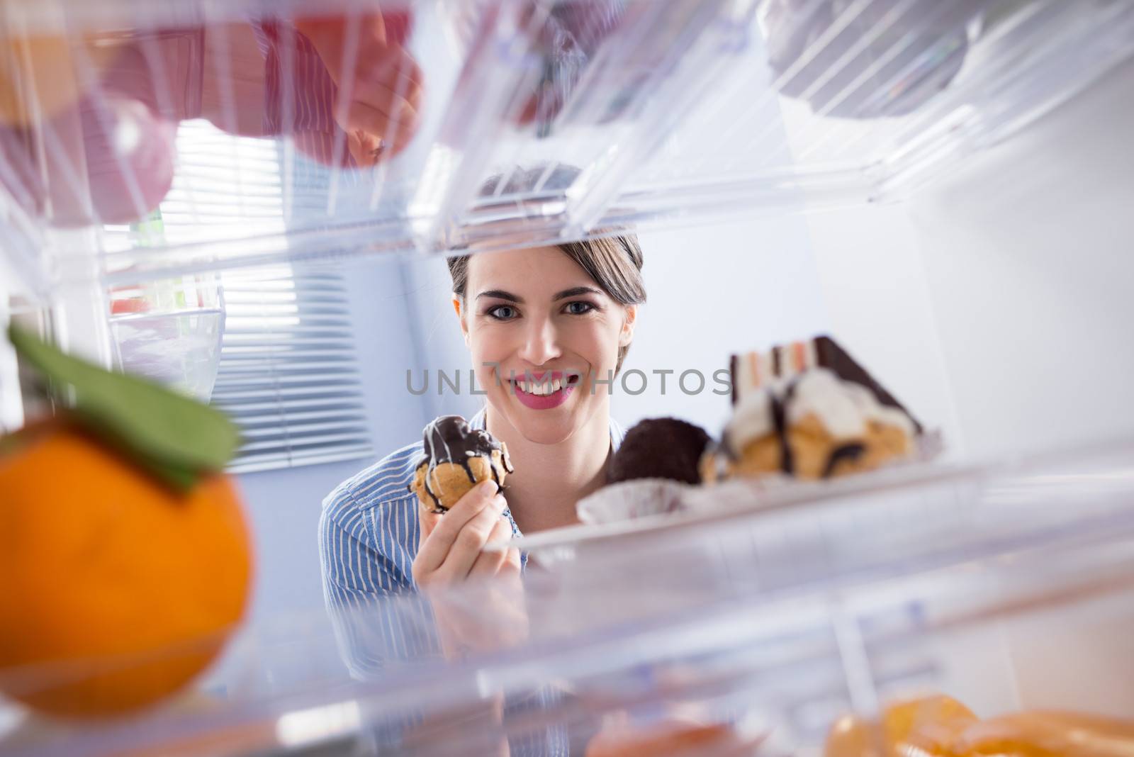 Young woman smiling and taking pastry with chocolate topping from fridge.