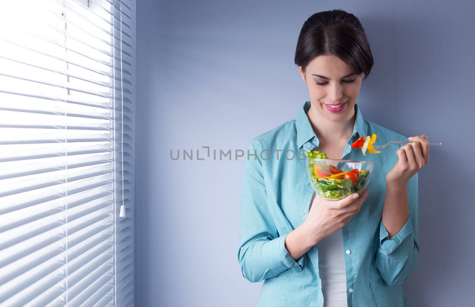 Woman smiling and eating salad in front of a window.