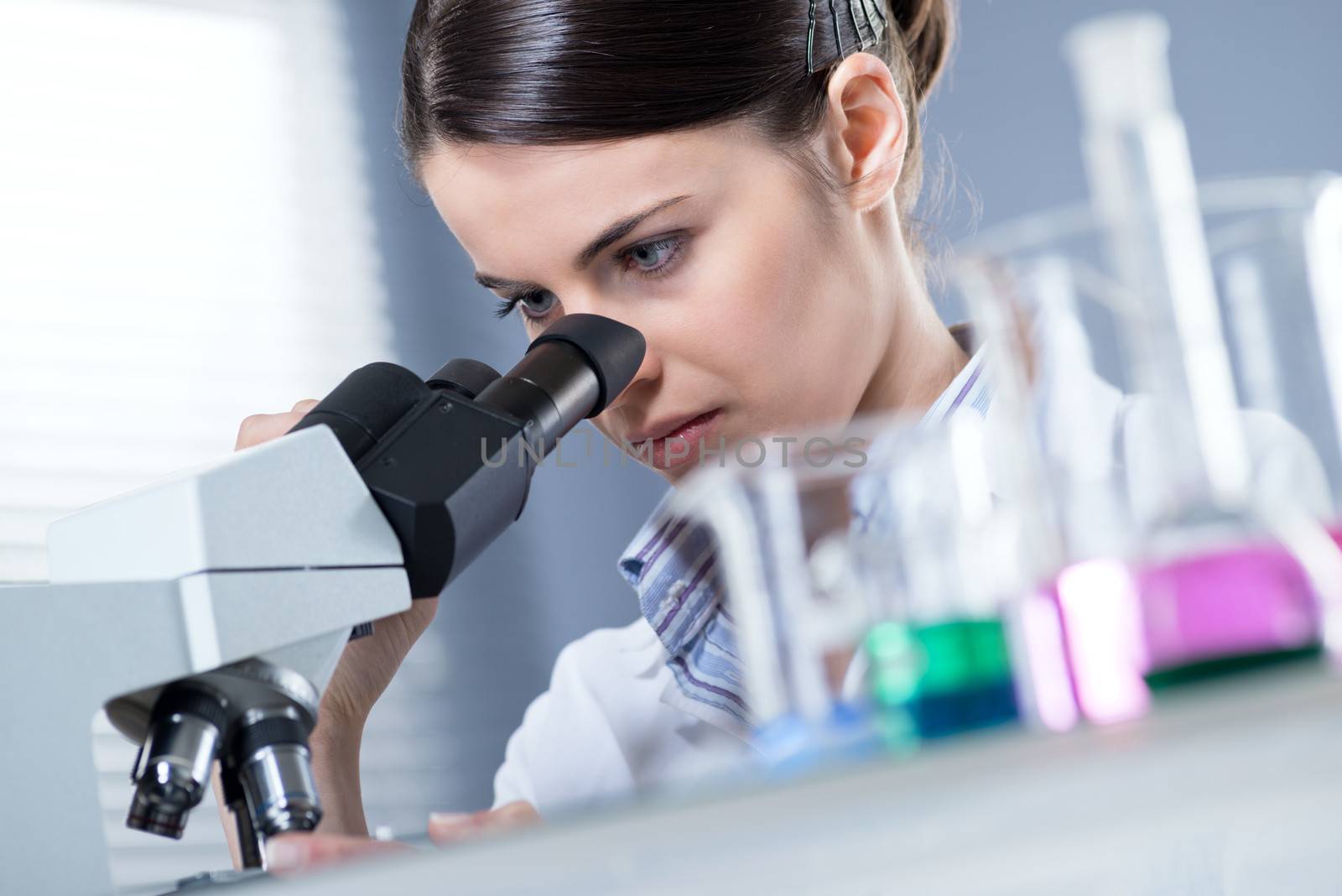 Young female researcher using microscope in the chemistry lab with laboratory glassware on foreground.