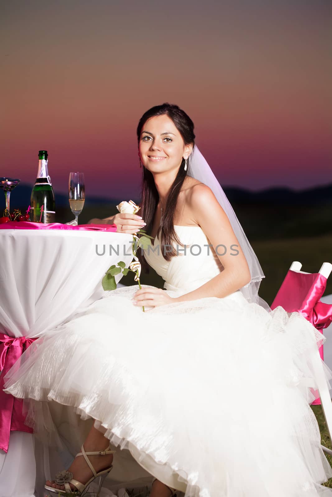 Bride is sitting at the wedding table outdoors with champagne and flower in the evening glow