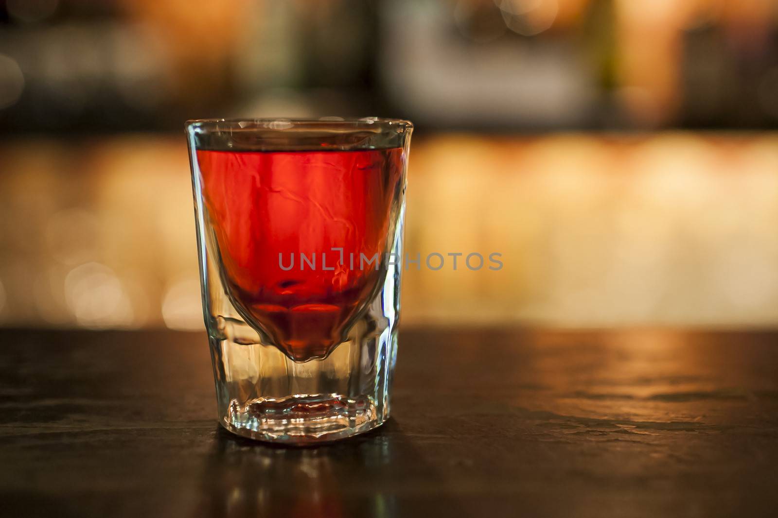 A glass of alcohol on the bar, vodka in the dark colour, red vodka, in the background the bar with bottles, shallow DOF.