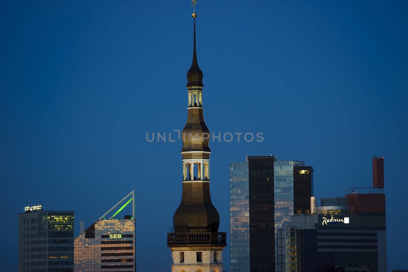 Photo presents fragment of the old town in Estonia, Talinn at night, visible illuminated church tower, modern office blocks in the background.