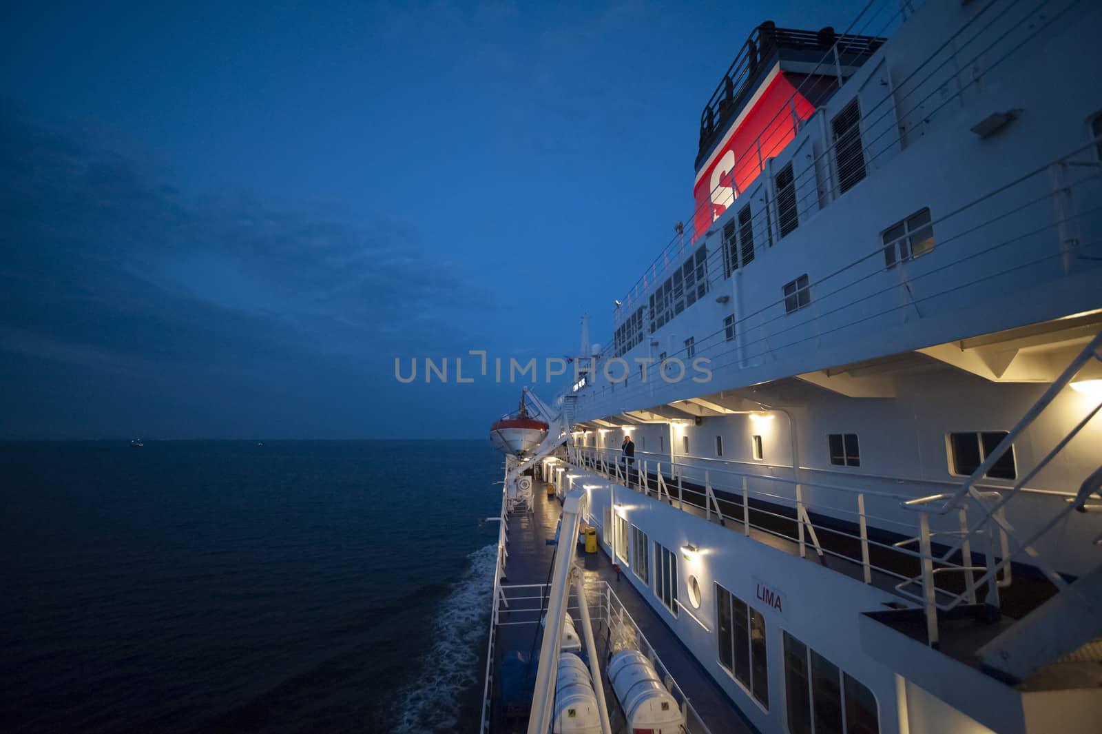 Photo presents one side of the big white modern ferry, with visible illuminate decks and a red funnel, navy blue night sky and calm sea in the background.