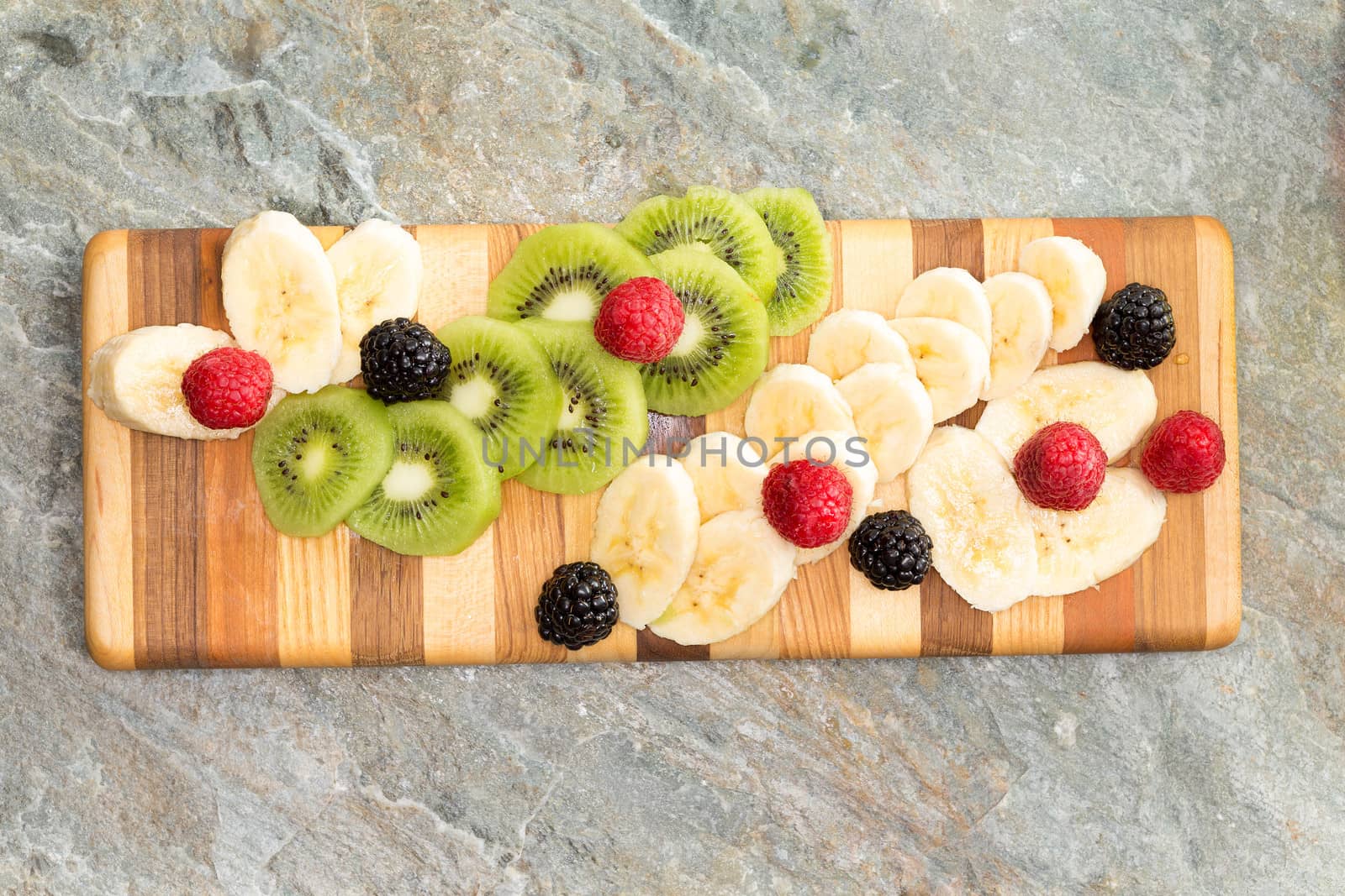 Sliced fresh ingredients ready for a fruit salad displayed on a decorative wooden striped chopping board including tropical kiwifruit, banana, raspberries and blackberries on a stone counter