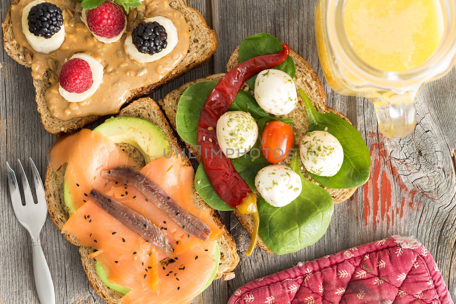 View from above of a selection of healthy picnic sandwiches topped with smoked salmon, anchovies, avocado, mozzarella, baby spinach, chili and fresh berries with a jug of orange juice