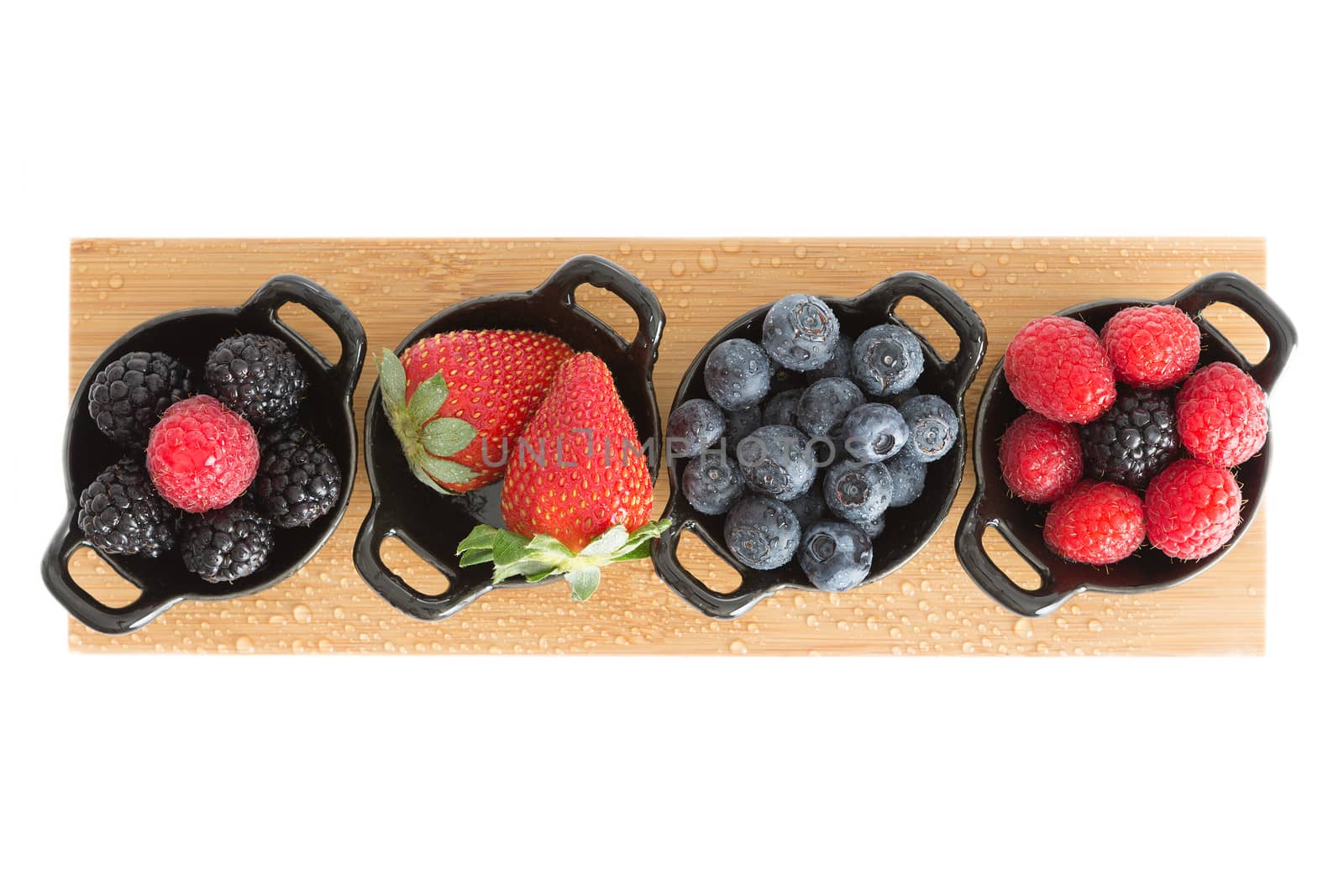 Healthy juicy autumn or fall strawberries, blueberries,. blackberries, and raspberries displayed in individual ceramic ramekins on a wooden board isolated on a white background, view from above