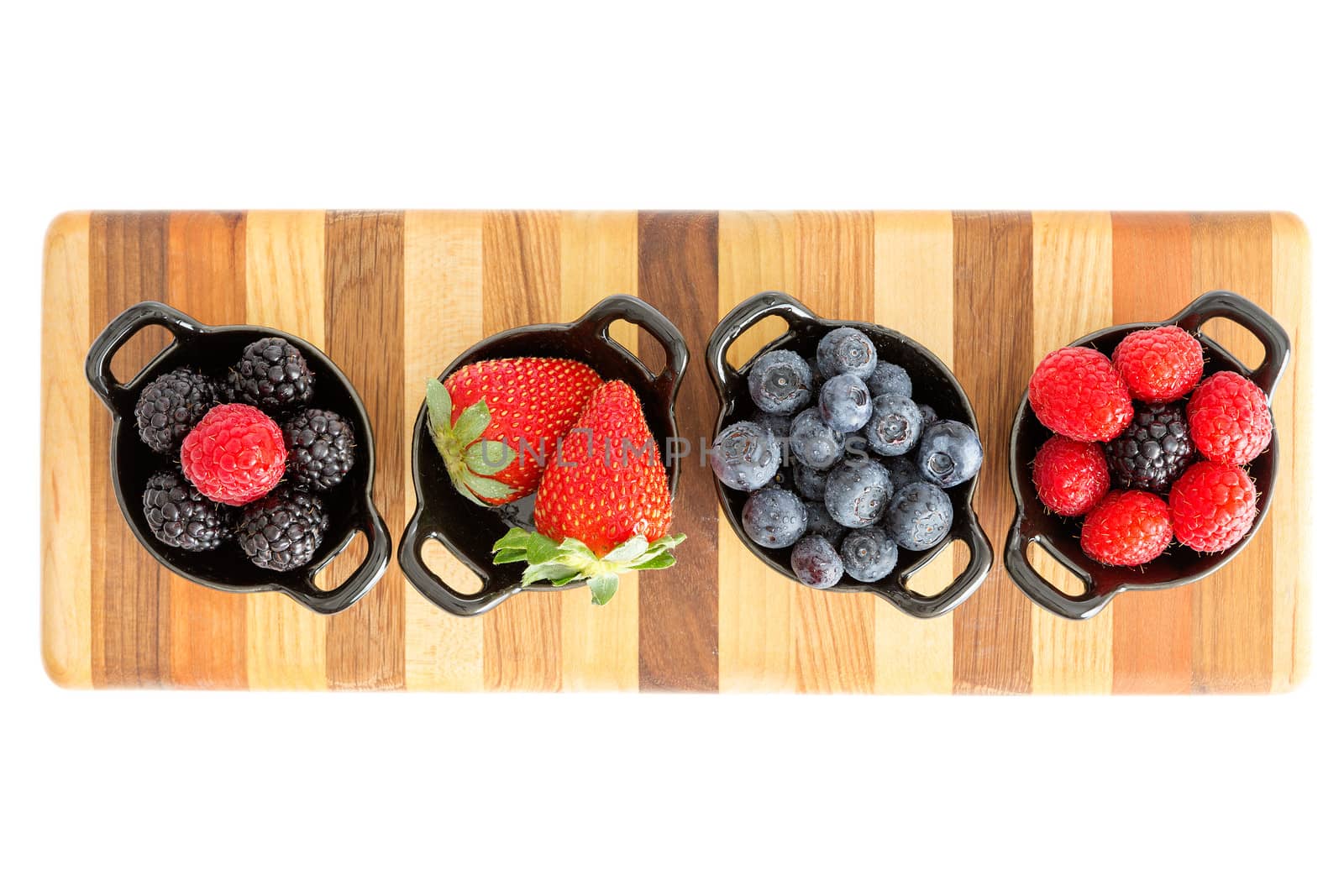 Selection of different ripe fresh autumn berries including strawberries, blueberries, blackberries and raspberries in separate dishes on a decorative striped wooden board, closeup overhead view