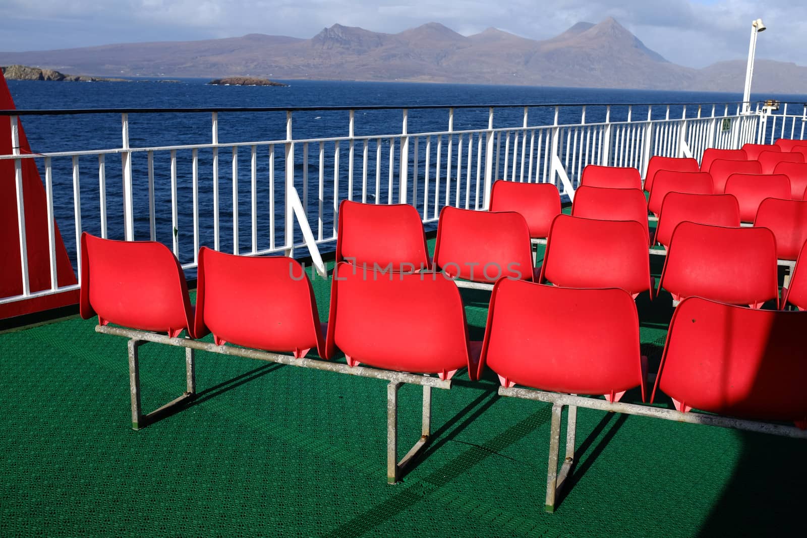 Rows of red plastic seating on a green rubber floor with white railings and a view of the sea and mountains in the distance.