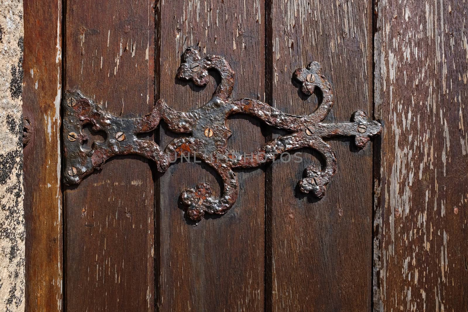 An ornate metal hinge with rust and flaking paint attached to a stained wooden door.