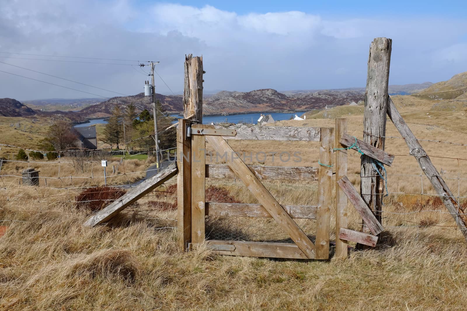 A wooden gateway with posts and a fence on moorland with a view to the sea and hills against a blue cloud sky.