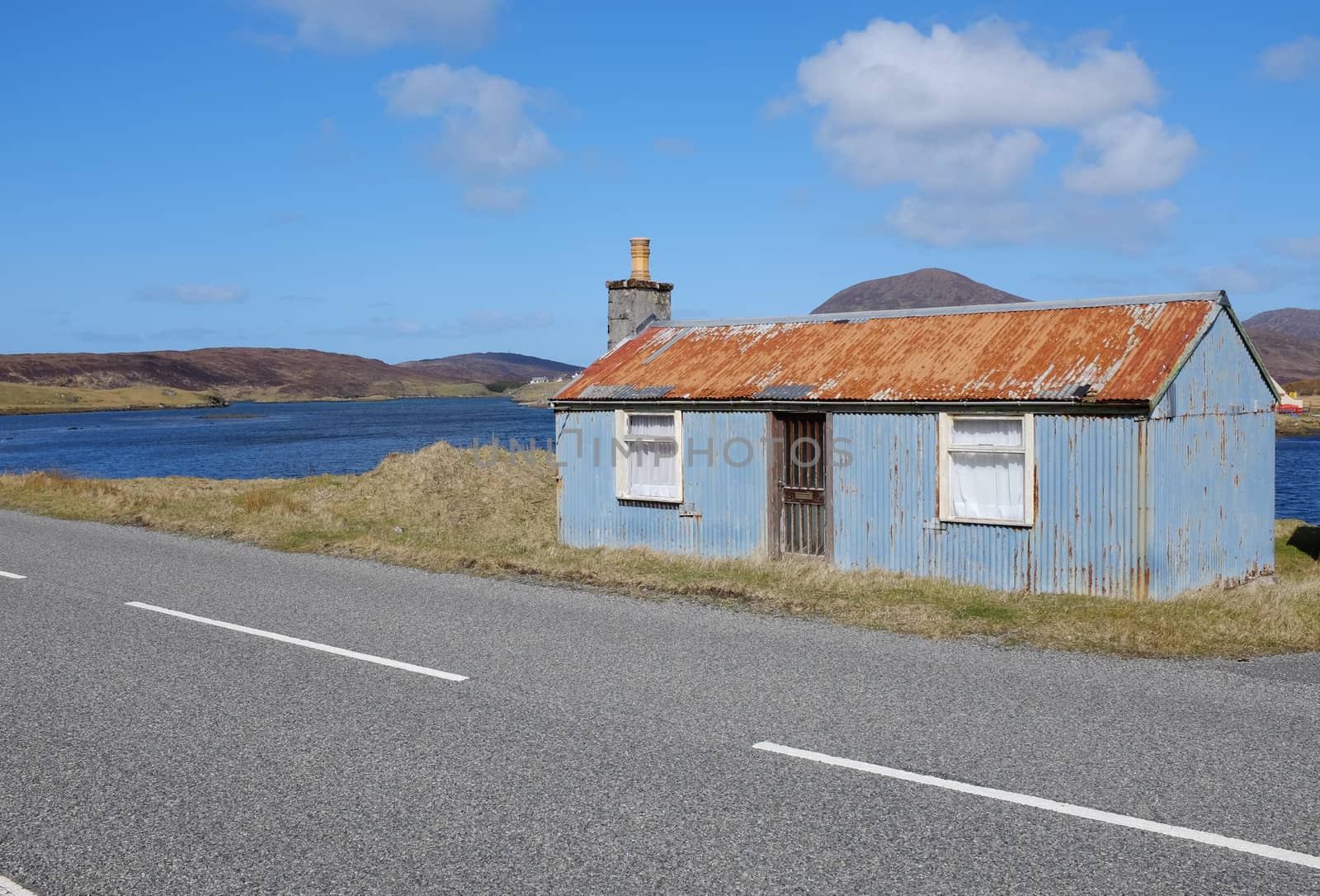 A derelict corrugated metal building on the side of the road with water and hills in the distance under a blue sky.