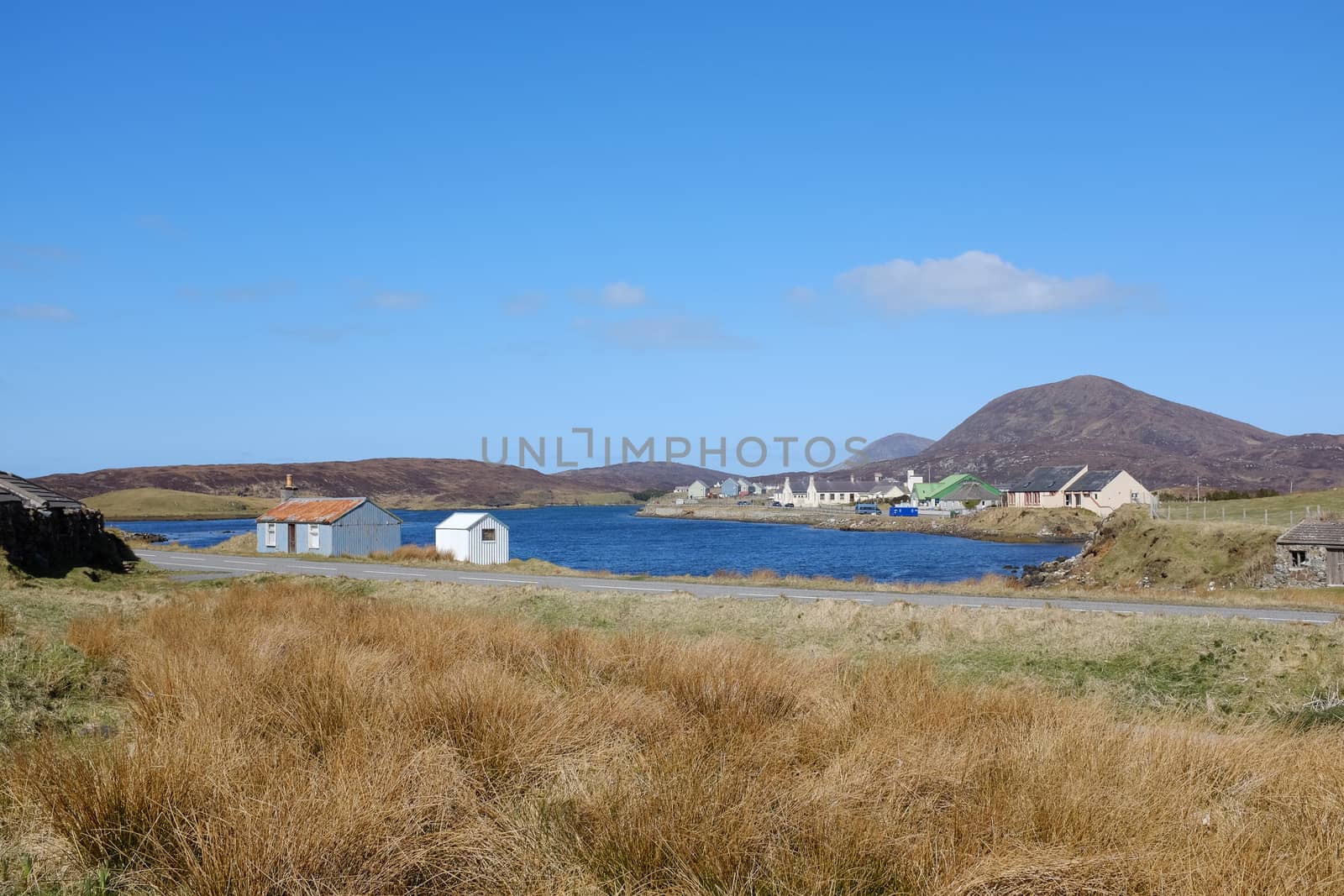 Grasses lead to tin huts overlooking a small loch with the village of Leverburgh, Isle of Harris, Scotland, UK.