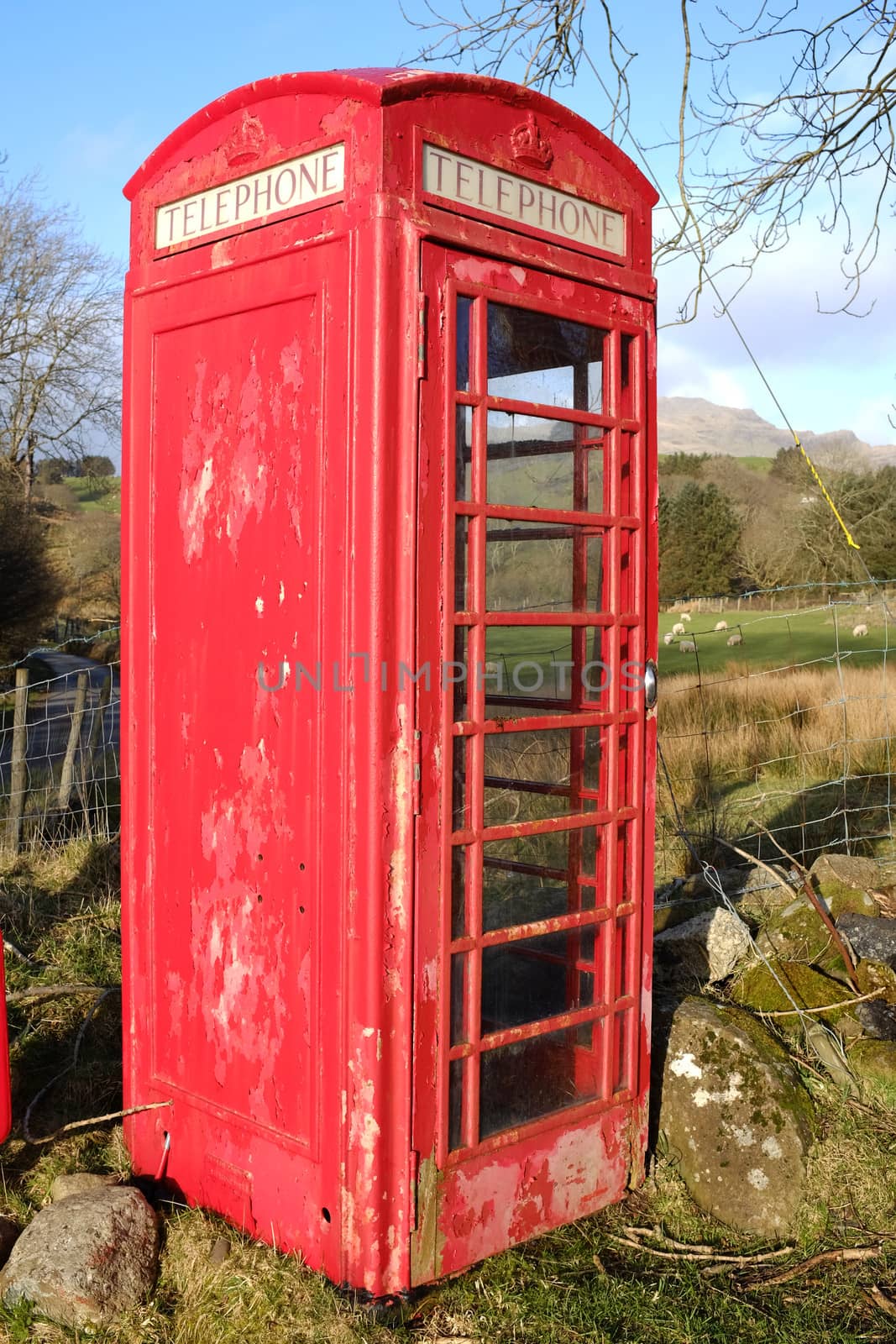 A vintage red British telephone box with flaking paint and 'telephone' sign in a rural setting.