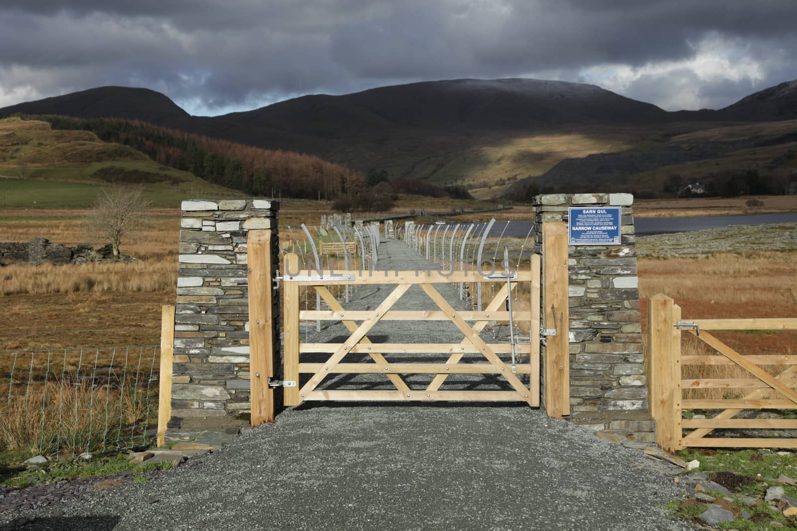 A wooden gate leads to a causeway with a hardcore path flanked by metal fencing with cloud covered mountains in the distance.