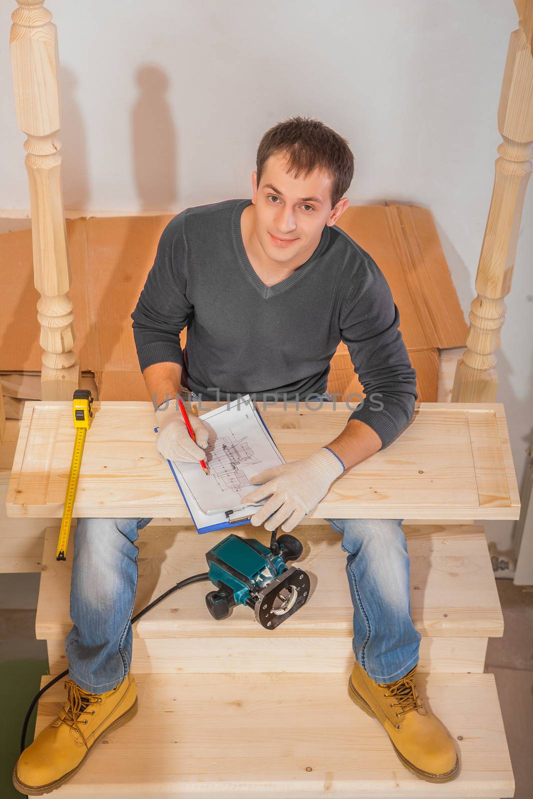 a young handsome man wearing working clothes sitting on ladder and work with blueprint