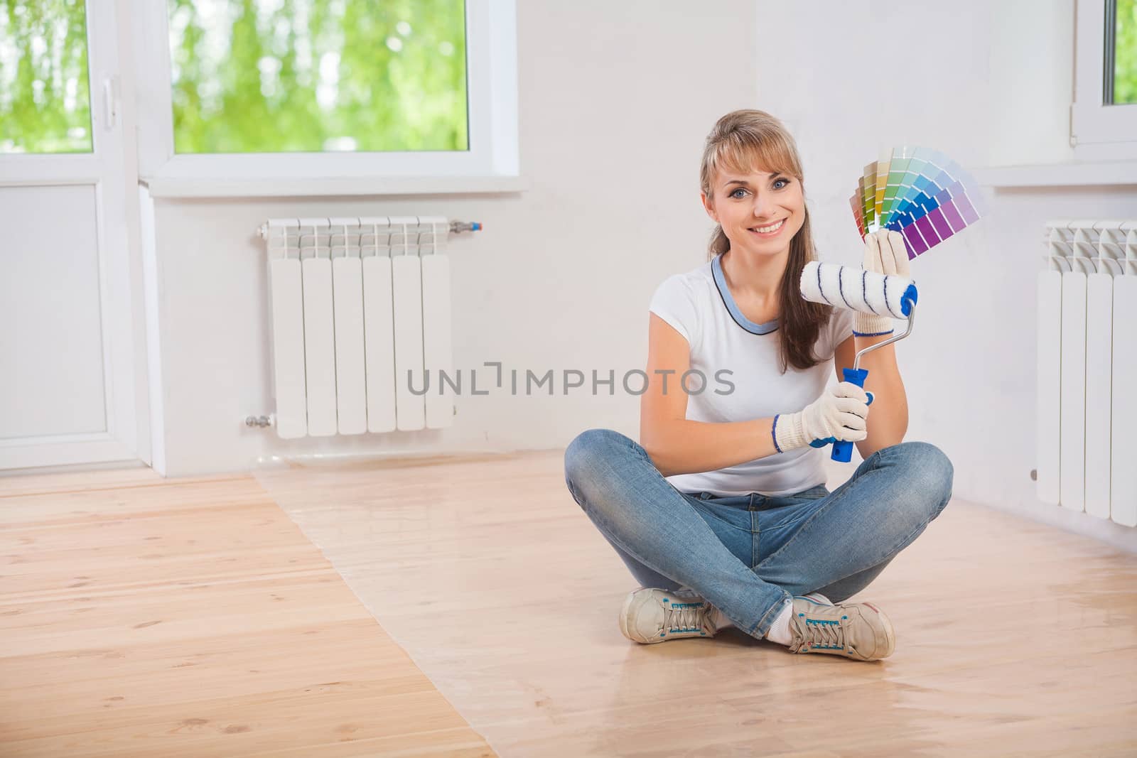 beautiful smiling female worker sitting on wooden floor and showing paintroller and color palette