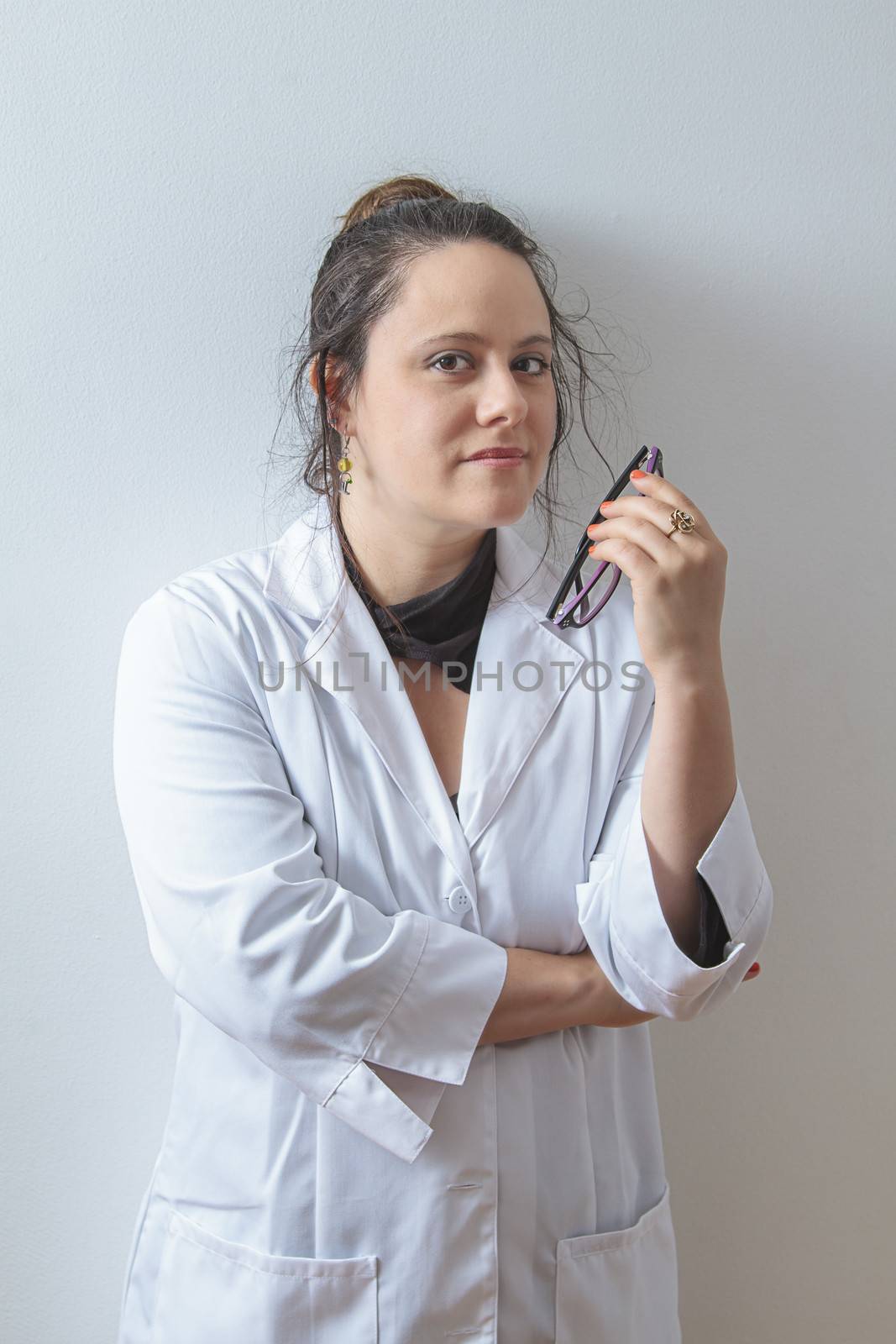 Woman doctor holding her glasses and leaning against a white wall