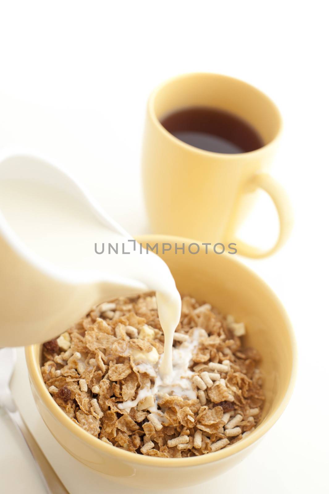 Pouring milk from a ceramic jug into a bowl of nutritional breakfast cereal or muesli with a freshly brewed mug of espresso coffee alongside