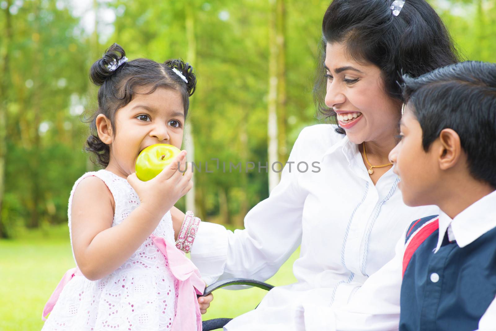 Happy Indian family. Asian girl eating an green apple at outdoor with mother and sibling.