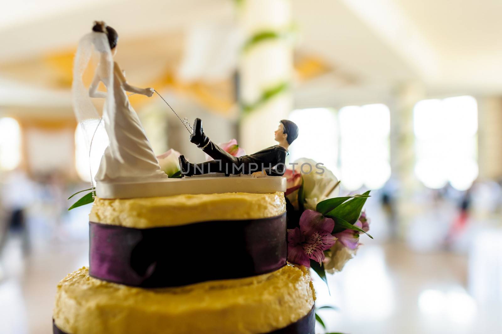 Fragment of yellow and purple wedding cake with funny decoration symbolizing groom pulled by leg on a leash by bride, wedding hall in background.