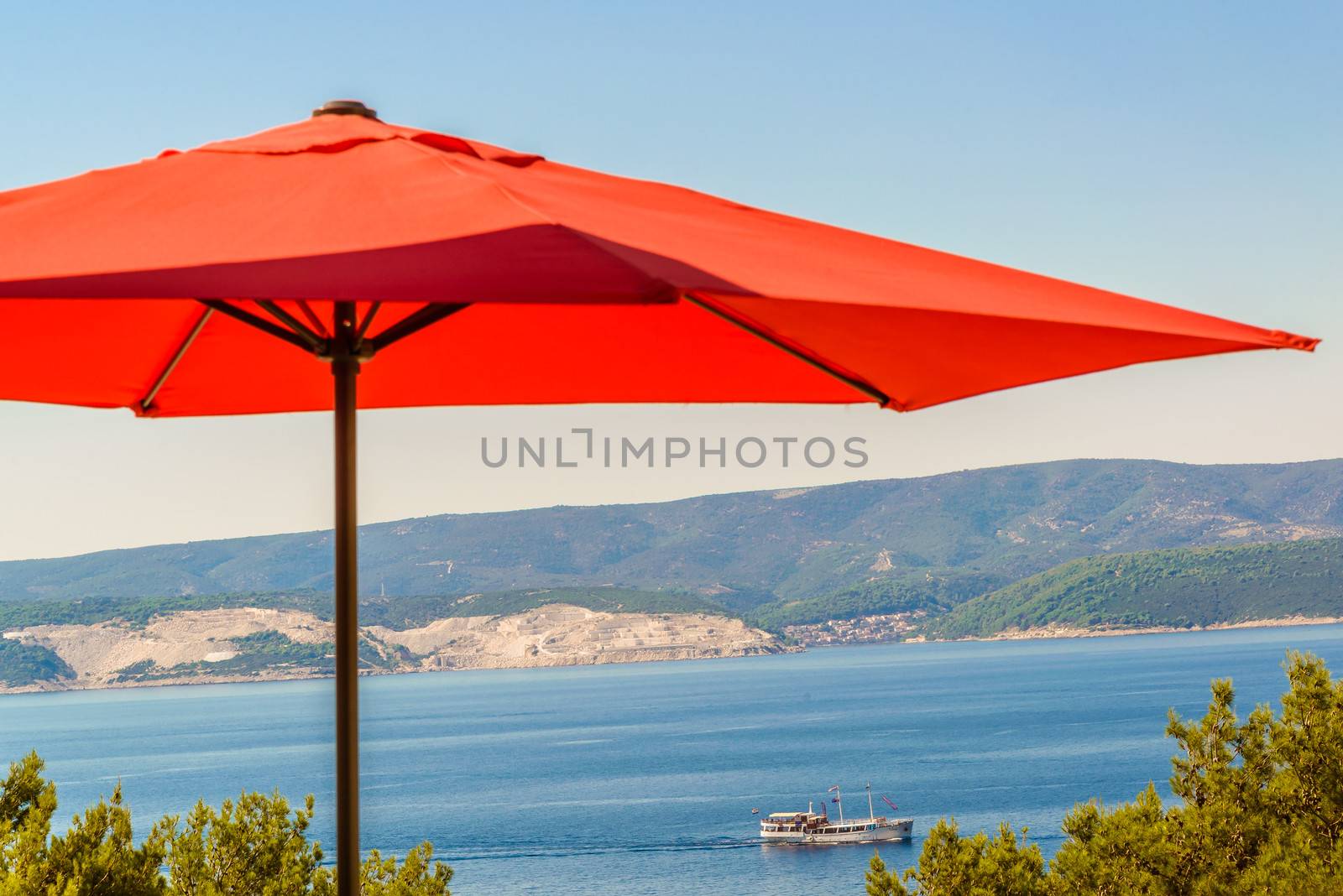 Red terrace, garden umbrella against blue sea and cloudless sky, visible coastline, ship in distance.