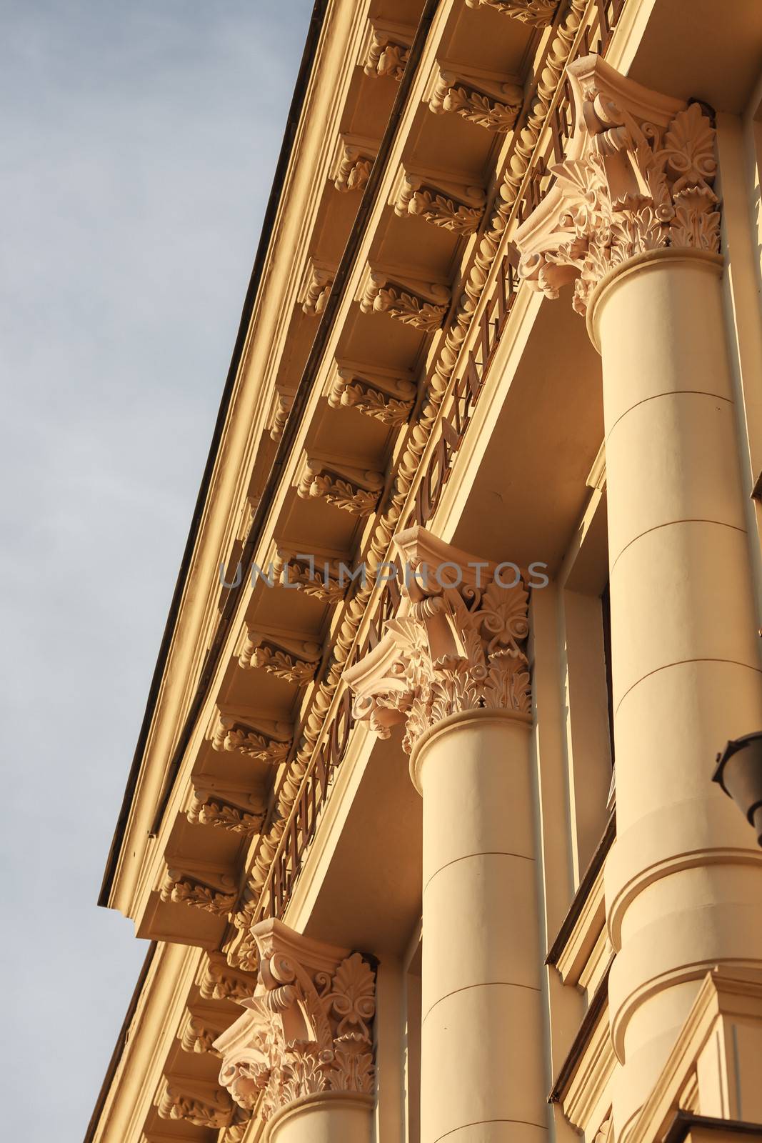 Fragment of classic or neoclassic building with columns and fine capitals, illuminated with warm summer sunlight, blue sky visible.